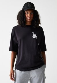 T-shirt en coton noir avec une coupe décontractée et des manches courtes, présentant un logo blanc sur la poitrine. Associé avec une casquette noire et un pantalon de jogging gris.