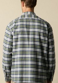 Uomo che indossa una camicia a quadri verde, blu navy e bianca a maniche lunghe, visto da dietro su uno sfondo beige uniforme.