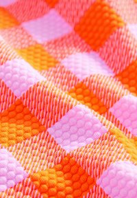 Textil mit einem erhöhten karierten Muster in Orange, Pink und Weiß. Die Textur ist taktil, mit wechselnden Farben, die visuelles Interesse erzeugen.
