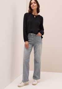 Blusa nera a maniche lunghe con un dettaglio a forma di chiave sul collo, abbinata a jeans grigi chiari a gamba larga e sneakers bianche su uno sfondo neutro.