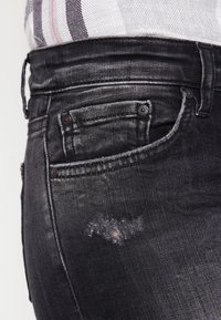 Jean en denim noir avec une texture délavée, des détails usés sur la cuisse, plusieurs poches et des boutons métalliques sur la ceinture.