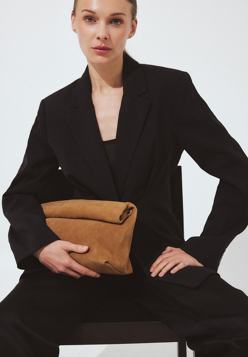 Woman in black suit seated, holding a large folded brown suede clutch bag against a plain white background.