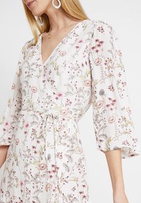 Floral-printed wrap dress in lightweight fabric, featuring a V-neck, elbow-length sleeves, and a tie at the waist. Pastel color palette.