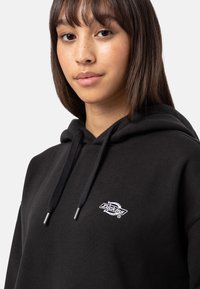 Black hoodie made of cotton with a relaxed fit. Features a drawstring hood and a small white logo on the chest. Smooth texture.