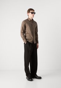 Camisa de manga longa castanha com colarinho, calças pretas e óculos de sol pretos. A camisa apresenta um corte ajustado e um design de botões.