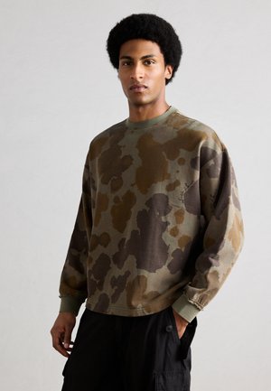 Mann mit Afro-Frisur, der ein Sweatshirt mit Tarnmuster und schwarze Cargohosen trägt, steht mit einer Hand in der Tasche vor einem einfarbigen Hintergrund.