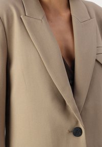 Blazer beige ajusté avec un col structuré, une fermeture à bouton noir unique et un tissu à texture lisse.