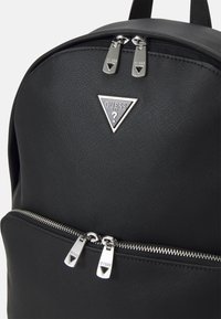 Crna ruksak s teksturiranim vanjskim dijelom, s prikazom srebrnog trokutastog logotipa i dvostrukim džepovima sa patentnim zatvaračem za pohranu. Akcenti uključuju srebrne metalne dijelove.
