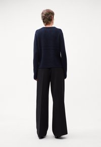 Tmavě modrý pletený svetr s kulatým výstřihem a žebrovanou strukturou, doplněný o široké černé kalhoty. Pohled zezadu zdůrazňuje celkový tvar postavy.