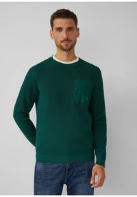 Groene gebreide trui met een textuur oppervlak, voorzien van een contrasterende groene zak en een witte kraag. Model draagt blauwe jeans.