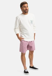 Biely mikina s zeleno-ružovým dizajnom na hrudi, skombinovaná s ružovými šortkami a čiernymi teniskami. Jednoduchý, neformálny outfit.