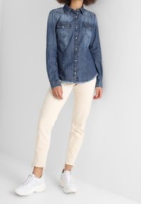 Dunkelblaue Jeanshemd mit Knöpfen und zwei Brusttaschen, getragen mit cremefarbenen Jeans und weißen Turnschuhen, mit ausgefransten Säumen.