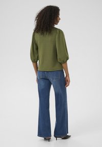 Haut vert texturé avec manches bouffantes associé à un jean bleu taille haute à jambes larges. Le modèle porte des talons noirs pointus.