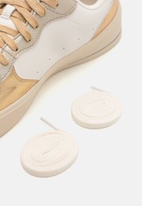 Valge ja kuldne sneaker tekstureeritud kangaga, millel on perforatsioonid ja heledavärvilised kingapaelad. Kaalub ka kaks lisaringi paela.