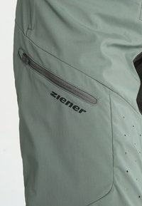 Gros plan sur un pantalon d'extérieur vert avec une poche zippée horizontale et le logo noir "ziener" imprimé en dessous de la poche.