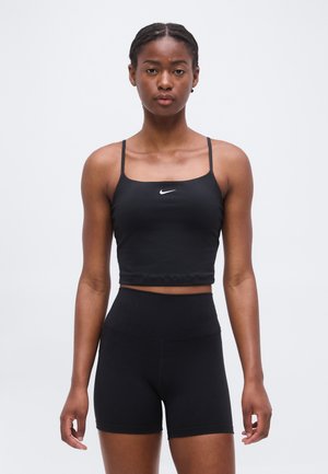 Atletyczna kobieta z warkoczami, ubrana w czarny krótki top Nike i wysokie spodenki rowerowe, stojąca na jednolitym białym tle.