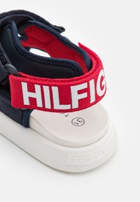Tummansininen sandaali, jossa on valkoinen kumipohja. Varustettu punaisella hihnalla, jossa lukee "HILFIGER" valkoisella tekstillä, yhdistettynä teksturoituun kankaaseen yläosassa.