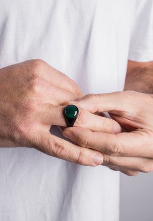 Black ring with a green stone, elongated oval shape, smooth surface, worn on a hand against a plain white background.