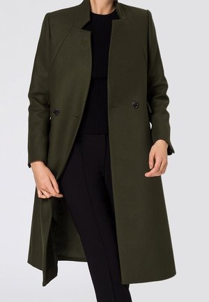Personne portant un long manteau vert foncé sur un haut noir et un pantalon noir, avec le manteau ouvert montrant la doublure intérieure.