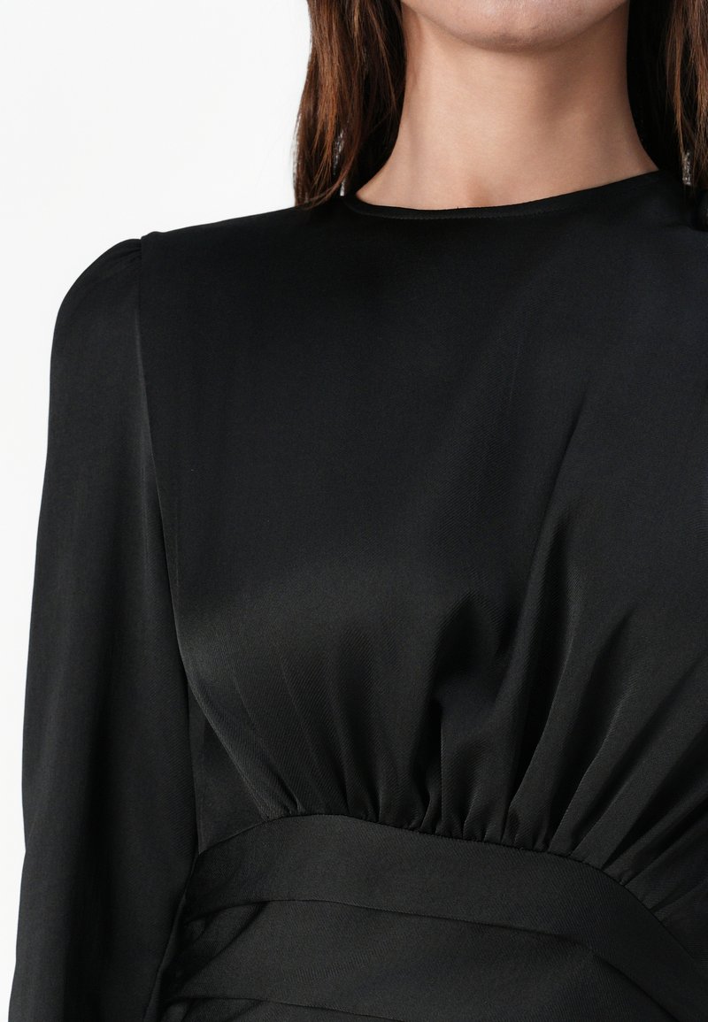 Schwarzes Kleid aus einem glatten, leichten Stoff mit einem gefalteten Taillendesign, langen Ärmeln und einem abgerundeten Ausschnitt.