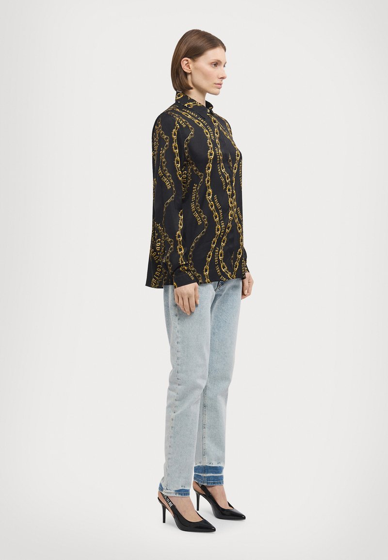 Camisa preta com um padrão de corrente, colarinho abotoado, mangas longas. Usada com jeans azul claro e sapatos de salto agudo pretos.