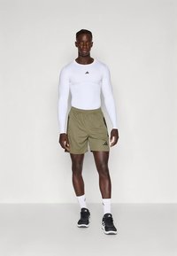 Bela športna majica z dolgimi rokavi, olivno zelene kratke hlače s črnimi poudarki, črni športni čevlji, bele nogavice in vidna Adidasova znamka.