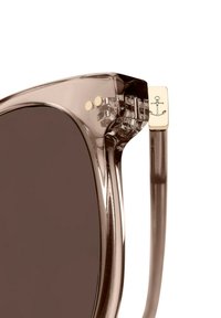 Ruskeasävyiset aurinkolasit, joissa on läpinäkyvät ruskeat kehykset. Niissä on kultaiset yksityiskohdat, joissa on ankkuri yksityiskohtana templeissä.