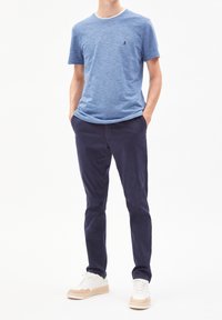 Muž oblečený v modrém melírovaném tričku s krátkým rukávem, tmavě modrých slim-fit kalhotách a bílých teniskách s béžovými a zelenými akcenty, stojící s rukama v kapsách.