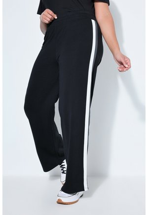 Person iført sorte bukser med brede ben og hvide striber på siden samt hvide sneakers med gummisåler, stående mod en hvid baggrund.