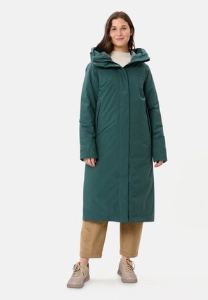 Femme portant un long manteau d'hiver à capuche vert foncé, un pantalon beige et des baskets beige clair, debout devant un fond clair uni.