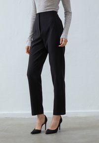 Pantalon noir taille haute avec plis à l'avant, coupe ajustée et poches ; associé à un haut gris à manches longues et des talons pointus noirs.