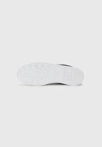 Crne slip-on cipele s teksturiranim gornjištem, s fleksibilnom bijelom gumom potplatom i okruglim uzorcima za prijanjanje.