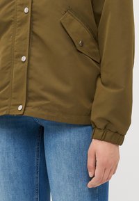 Olivenfarget jakke med en jevn, matt finish, med trykknapper, en sidelomme og elastiske mansjetter. Bært over lyseblå jeans.