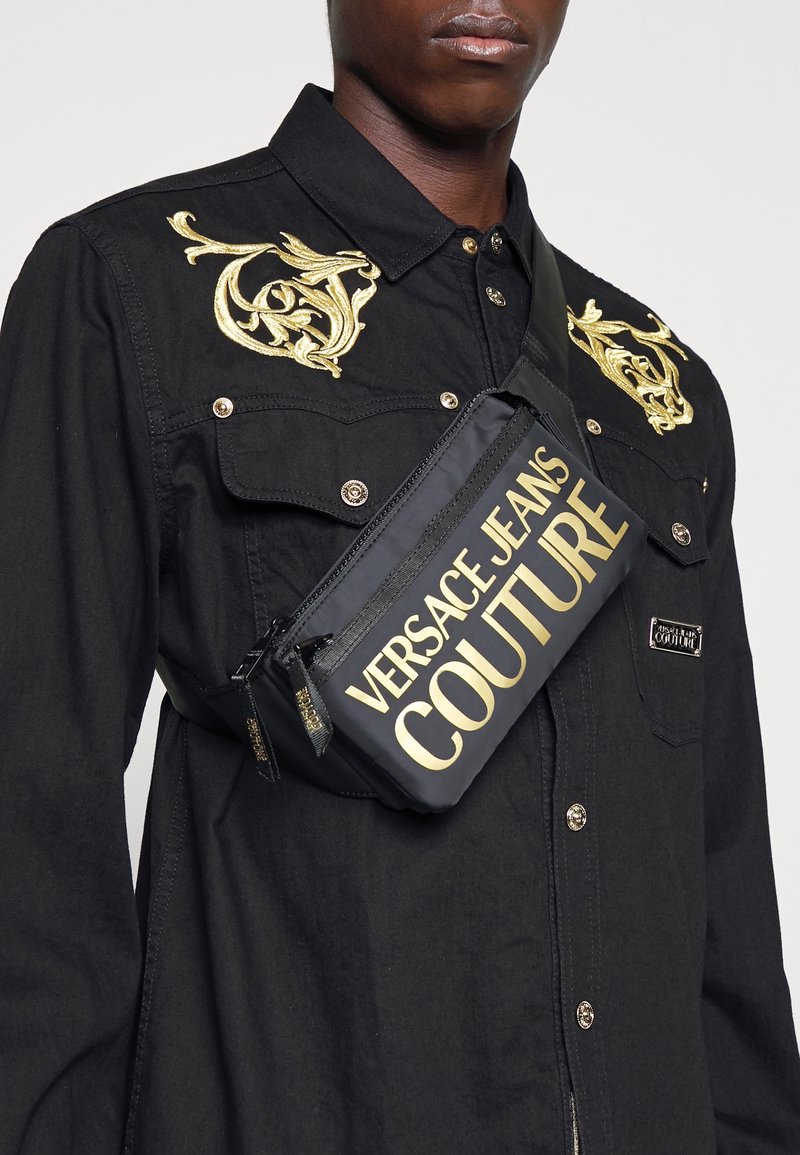 Personne portant une chemise noire avec une broderie dorée et un sac bandoulière noir avec "Versace Jeans Couture" en grandes lettres dorées.