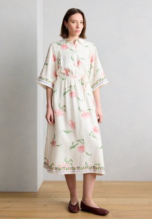 Žena stojaca v interiéri, oblečená v bielych midi šatách s ružovými kvetinovými vzormi, s tromi štvrťovými rukávmi, zaväzovaným pásom a bordovými balerínami.