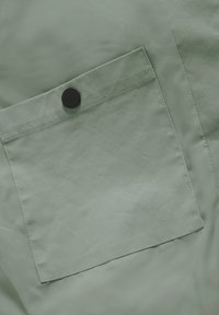 Světlězelená tkanina s hladkou texturou obsahuje obdélníkovou náprsní kapsu zabezpečenou černým patentním knoflíkem.