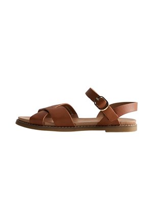 Brun læder flad sandal med krydsede stropper, ankrem med rund metalspænde og en beige gummisål.