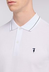 Bílá polo košile vyrobená z texturované tkaniny, s černě prošitým límcem a malým námořnickým logem na levé části hrudníku.