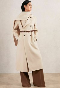 Beige wollen jas met een dubbelgeknoopte ontwerp, grote revers en een bruine leren riem. Gestyled met brede bruine broeken, met een zachte textuur.