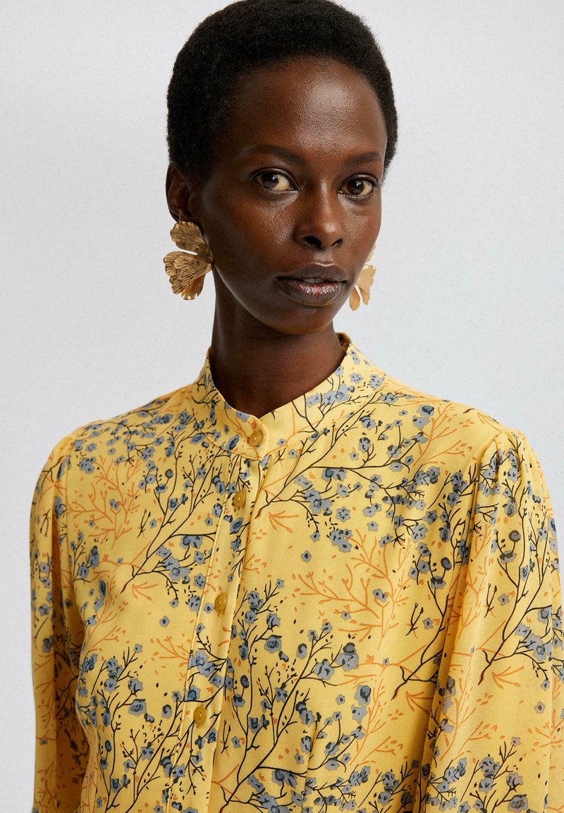 Gelbe Bluse mit floralem Muster in Blau und orangefarbenen Zweigen, Knopfleiste vorne und Stehkragen. Große, dekorative Ohrringe.