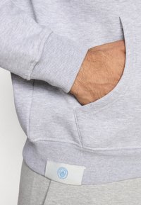 Γκρίζα φούτερ με ριμπ μανσέτες και πλευρική τσέπη, με ένα μικρό μπλε λογότυπο κοντά στο στρίφωμα. Μαλακό, υφασμένο ύφασμα.