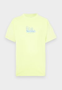 Neónovo žlté bavlnené tričko s krátkymi rukávmi, s modrým vyšívaným logom „Café Swoosh“ na prednej strane. Hladká textúra.