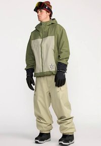 Outdoor-Jacke in Olivgrün mit beigem Vorderteil, mit Kapuze. Kombiniert mit beigen Hosen und schwarzen Handschuhen, getragen mit schwarzen Stiefeln.