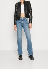 Zwarte cropped leren jas met hoge kraag, gecombineerd met lichtblauwe uitlopende spijkerbroek en zwarte laarzen. Minimalistisch ontwerp met ritssluitingen.