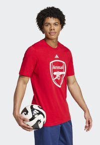 Červené tričko s krátkym rukávom s bielym logom Arsenal. Model drží čiernobiely futbalový loptu, v páre s modrými šortkami.