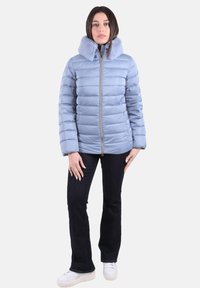 Chaqueta acolchada azul claro con capucha forrada de piel sintética, cierre de cremallera y acolchado horizontal; combinada con vaqueros oscuros y zapatillas blancas.