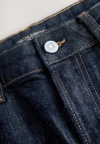 Jeans de mezclilla oscura con cierre de botones y costuras en contraste de color naranja. La etiqueta de la marca, "Tabitha Simmons", es visible cerca de la cintura.