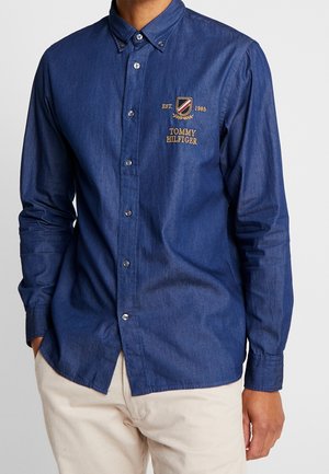 Mężczyzna noszący niebieską koszulę na długi rękaw z logo "Tommy Hilfiger" i beżowe spodnie, stojący z rękami w kieszeniach.