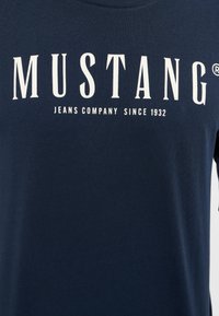 Mörkblå bomulls-T-shirt med vit tryckt text "MUSTANG JEANS COMPANY SINCE 1932." Rund halsringning, korta ärmar, slät textur.