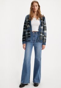 Noor noor seljas sinine ruuduline kardigan, valge lühikese varrukaga topp, kõrge vöökohaga figureerivad teksad ja mustad paksud kingad, seismas valge tausta vastas.